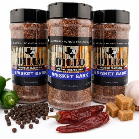 Three 9 oz bottles of Dillo Brisket Bark Premium Seasoning value bundle surrounded by fresh ingredients including jalapeños, black peppercorns, dried red chilis, garlic, and brown sugar cubes.
